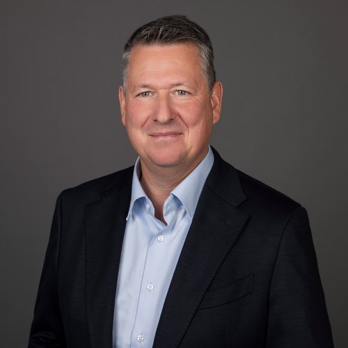 Martin Westhauser im Portrait Porträt eines mittelalten Mannes in dunklem Anzug und hellblauem Hemd vor neutralem Hintergrund, lächelnd und direkt in die Kamera blickend.