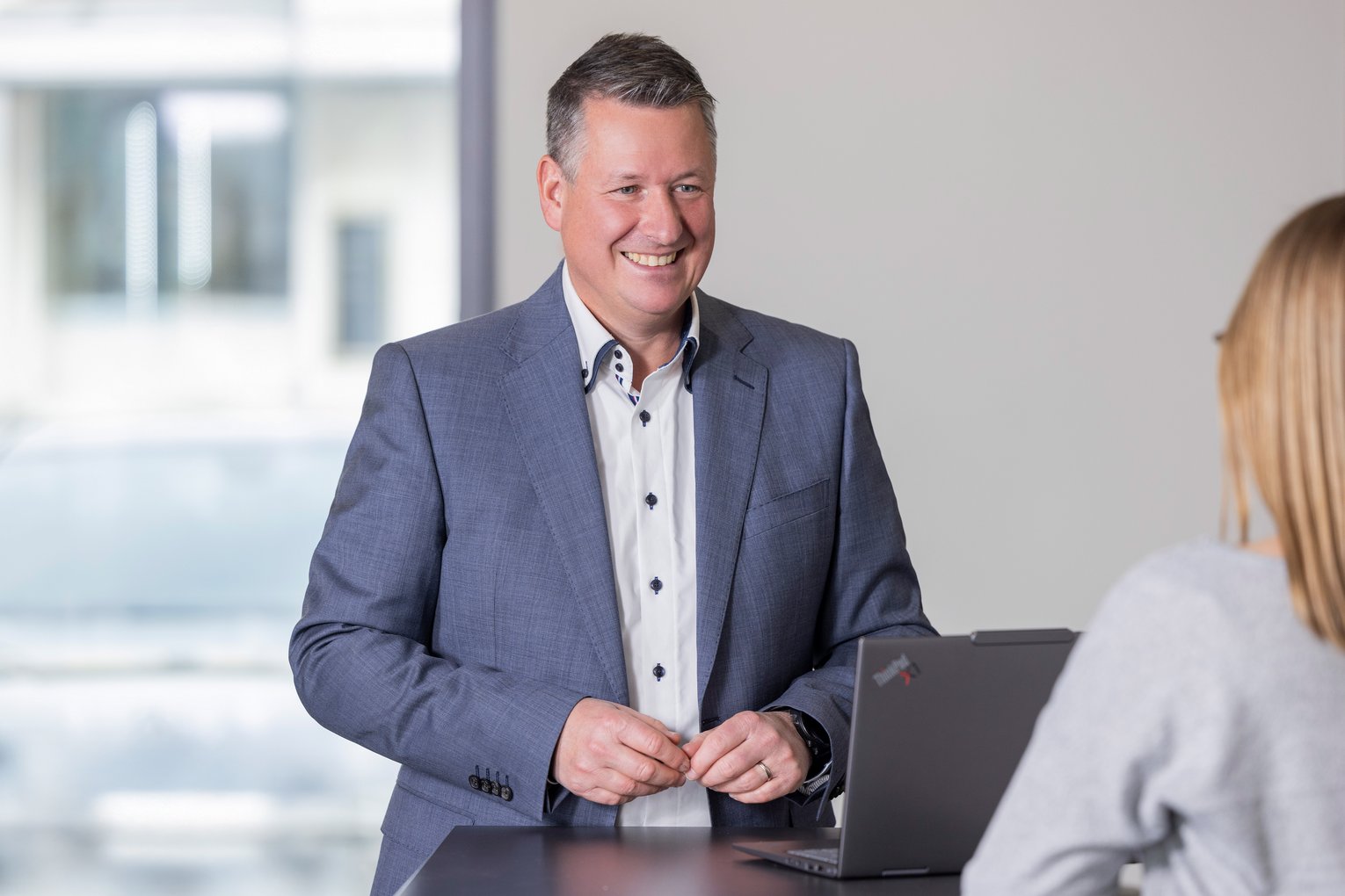 Martin Westhauser im Gespräch Ein Mann im Anzug steht lächelnd an einem Tisch und spricht mit einer Person, die vor einem Laptop sitzt.