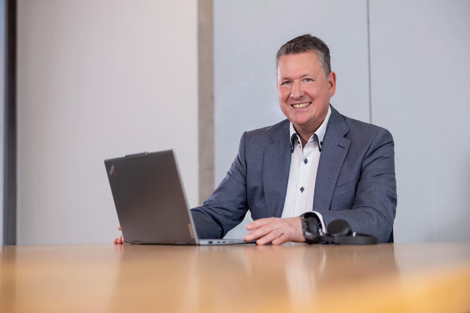 Martin Westhauser bei der Arbeit Ein lächelnder Mann im Anzug sitzt an einem Tisch und arbeitet an einem aufgeklappten Laptop. Neben ihm liegt eine Brille.